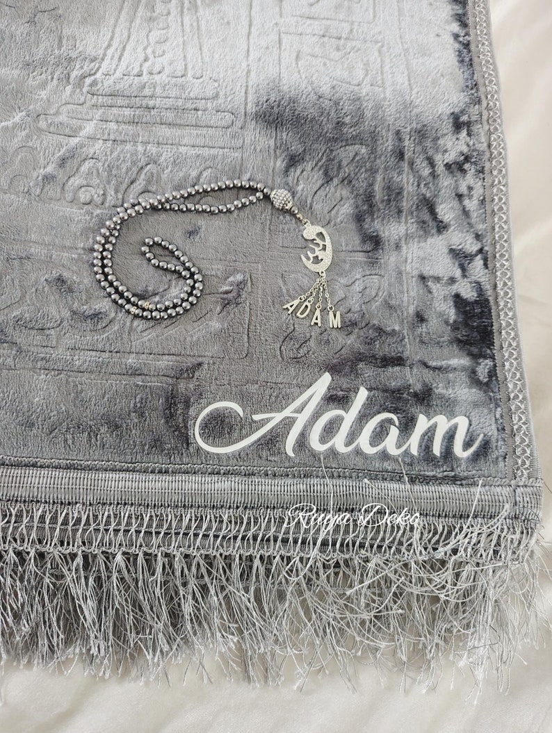 May include: A gray prayer rug with a silver tassel and a silver prayer bead necklace with a crescent moon charm. The rug has the name "Adam" embroidered on it in white.
