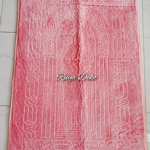 May include: A pink prayer rug with a white intricate design. The rug has a fringe border and the text "Ruyya Deko" is printed in the center.