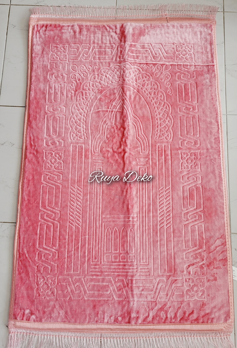 May include: A pink prayer rug with a white intricate design. The rug has a fringe border and the text "Ruyya Deko" is printed in the center.