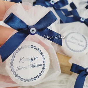 May include: White favor bags with blue ribbon ties and blue evil eye charms. The bags contain white round favors with blue evil eye designs and the text "Koray'in Sunnet Mevlidi".