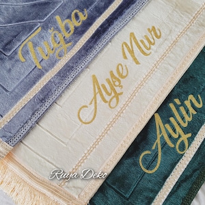 May include: Three prayer rugs with gold embroidery. The top rug is gray with the name "Tugba" embroidered on it. The middle rug is cream with the name "Ayse Nur" embroidered on it. The bottom rug is green with the name "Aylin" embroidered on it.