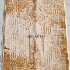 May include: A beige prayer rug with intricate embossed designs, including an archway and geometric patterns. The rug has a gold fringe border and the text "Ruyya Deko" is visible in the center.