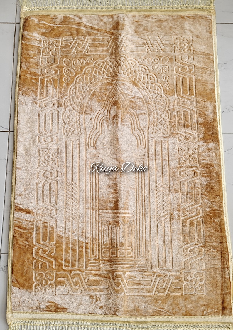 May include: A beige prayer rug with intricate embossed designs, including an archway and geometric patterns. The rug has a gold fringe border and the text "Ruyya Deko" is visible in the center.