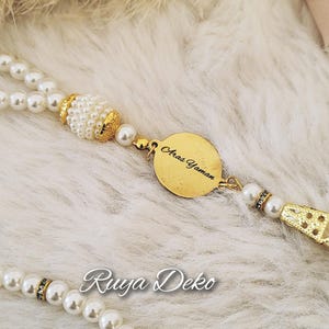 Könnte beinhalten: Eine Perlenkette mit einem goldenen Anhänger mit dem Namen "Aras Yaman". Die Halskette hat einen dekorativen Abschnitt aus Gold- und Perlenperlen und den Text "Ruga Deko".