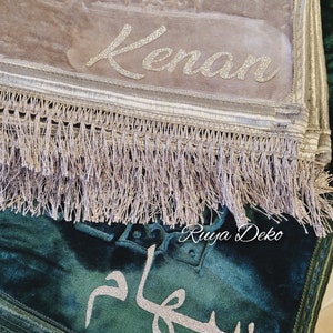 May include: Two prayer rugs with fringed edges. The top rug is beige with the name "Kenan" embroidered in silver glitter. The bottom rug is dark green with the word "Salam" embroidered in silver glitter. The bottom rug has the text "Ruyya Deko" in the corner.