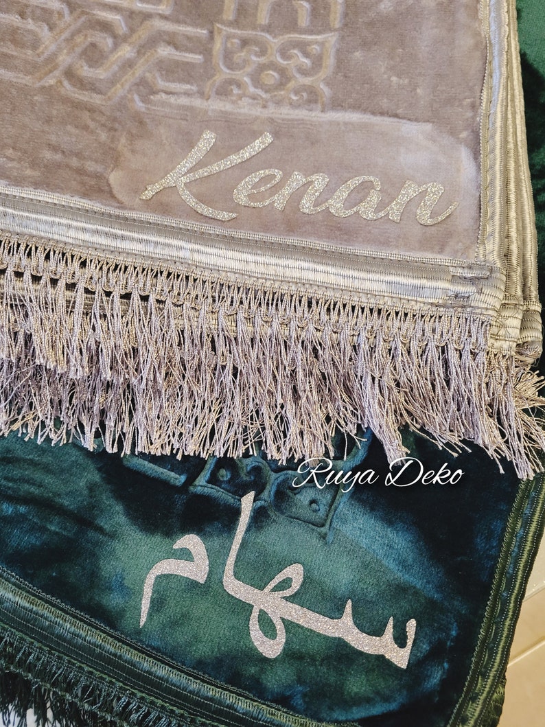 May include: Two prayer rugs with fringed edges. The top rug is beige with the name "Kenan" embroidered in silver glitter. The bottom rug is dark green with the word "Salam" embroidered in silver glitter. The bottom rug has the text "Ruyya Deko" in the corner.