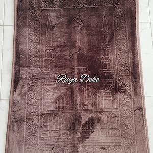 May include: A brown prayer rug with a detailed embroidered design of a mosque. The rug has a fringe border and the words "Ruyya Deko" are printed in the center.