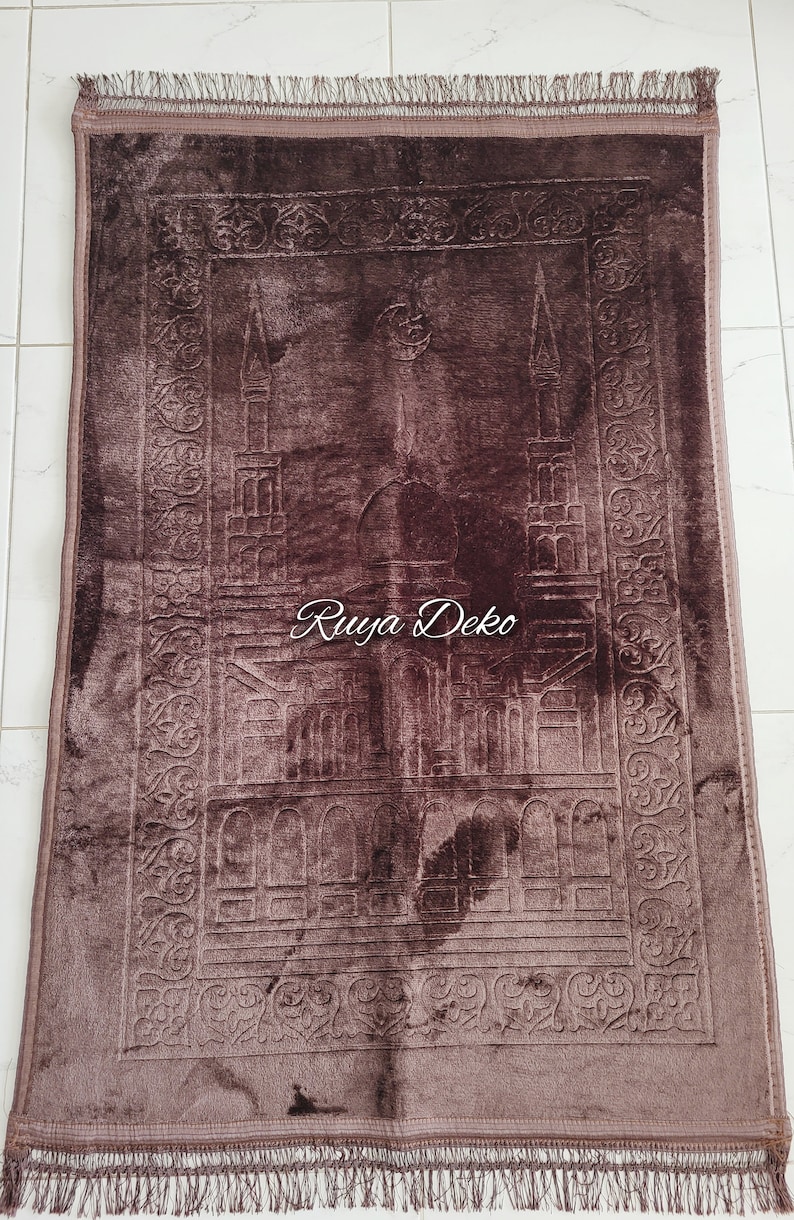 May include: A brown prayer rug with a detailed embroidered design of a mosque. The rug has a fringe border and the words "Ruyya Deko" are printed in the center.