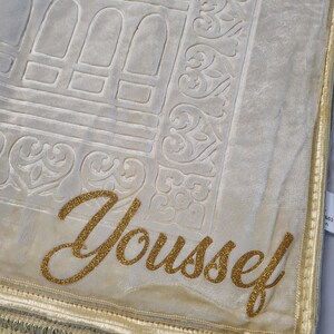 May include: A cream-colored prayer rug with a gold embroidered border and the name "Youssef" in gold glitter lettering.