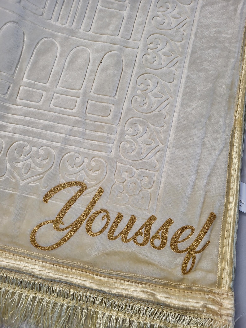 May include: A cream-colored prayer rug with a gold embroidered border and the name "Youssef" in gold glitter lettering.