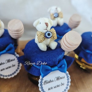 Pode incluir: Potes de lembrancinhas azuis e brancos com um urso de pelúcia branco vestindo uma gravata borboleta azul e dourada e um amuleto azul de olho grego. Os potes têm uma fita azul e um mexedor de mel de madeira. O rótulo no pote diz "Biska Miras ser xere be".