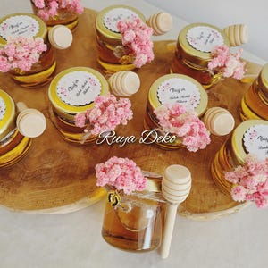 Könnte beinhalten: Kleine Gläser gefüllt mit Honig, bedeckt mit goldenen Deckeln, rosa Blüten und hölzernen Honiglöffeln. Jedes Glas hat ein Etikett mit Text. Die Gläser sind auf einer Holzoberfläche angeordnet.