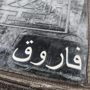 May include: A gray prayer rug with a black border and fringe. The rug has a geometric pattern and the word "فَارُوق" in white lettering.