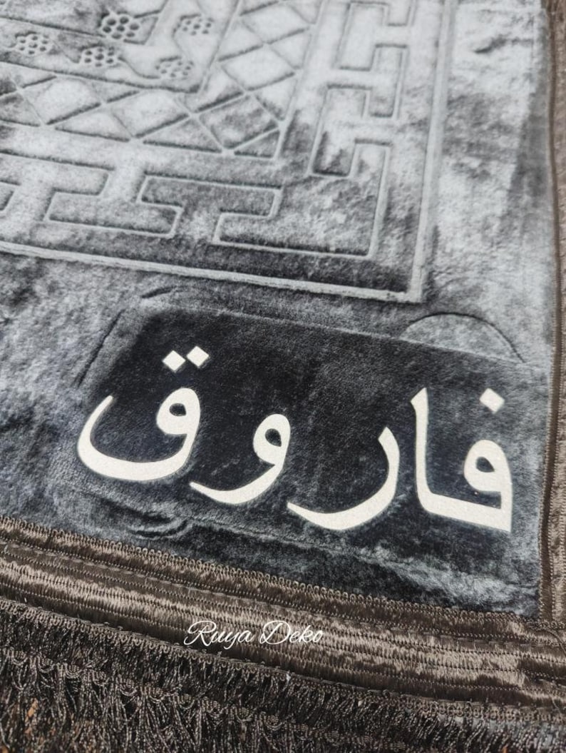 May include: A gray prayer rug with a black border and fringe. The rug has a geometric pattern and the word "فَارُوق" in white lettering.
