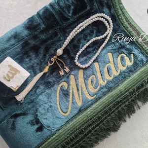 K&ouml;nnte beinhalten: Ein gr&uuml;ner Gebetsteppich mit goldener Stickerei, auf dem "Melda" steht. Der Teppich hat einen gr&uuml;nen Fransenrand und wird mit einer wei&szlig;en Gebetsmatte, einer wei&szlig;en Gebetsperlenkette und einem kleinen wei&szlig;en Quadrat mit arabischer Kalligraphie geliefert.