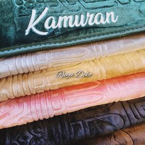 May include: A stack of prayer rugs in various colors, including green, beige, pink, black, and brown. The top rug is green and has the word "Kamuran" embroidered on it. The bottom rug is brown.