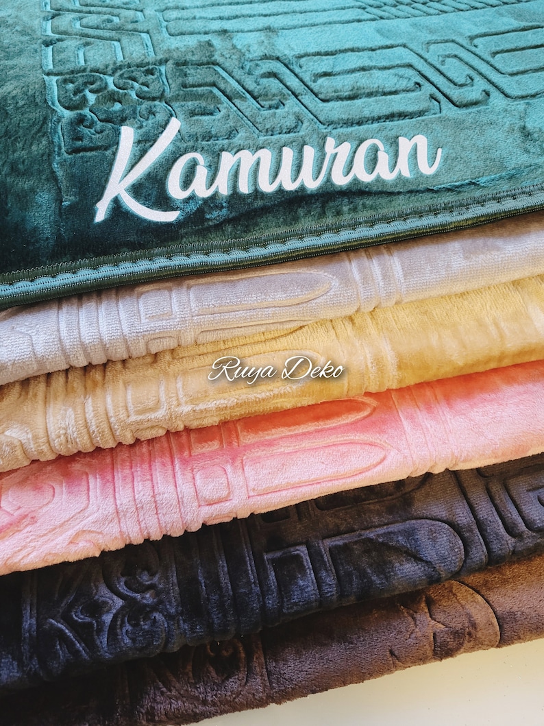 May include: A stack of prayer rugs in various colors, including green, beige, pink, black, and brown. The top rug is green and has the word "Kamuran" embroidered on it. The bottom rug is brown.
