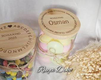 May include: Two glass jars with wooden lids, filled with colorful candies. The lids are engraved with text and a heart symbol. One jar says "Schulkind 2025". The other says "Osman". The jars are tied with twine.