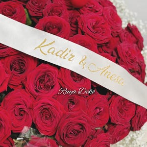 Könnte beinhalten: Ein Strauß roter Rosen mit einem weißen Satinband, das um sie herum gebunden ist. Das Band trägt den Text "Kadir & Aresa" in goldenen Glitzerbuchstaben. Der Text "Ruyua Deko" befindet sich ebenfalls auf dem Band.