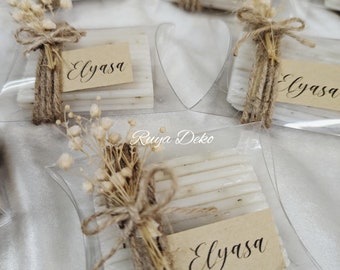May include: A collection of small, white, rectangular soaps wrapped in clear plastic with a brown twine bow and dried white flowers. Each soap has a small, brown paper tag with the name "Elyasa" written in black calligraphy.