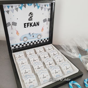 Op de afbeelding: Een zwarte doos met een wit deksel met kleine witte doosjes met een blauwe auto en de naam "EFKAN" en het nummer "2" erop. Het deksel heeft een blauwe auto en de tekst "2 EFKAN" erop. De doos is omringd door snoepstokken en doorzichtige plastic zakken.