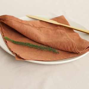 May include: A folded terracotta linen napkin on a white plate with a gold knife and a sprig of greenery.