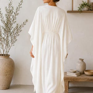 May include: A flowing, white caftan dress with a cinched waist. The dress has wide sleeves and a V-neck. The model is wearing sunglasses and has their hair up. The background includes a neutral-toned room with a wooden shelf and a large vase.