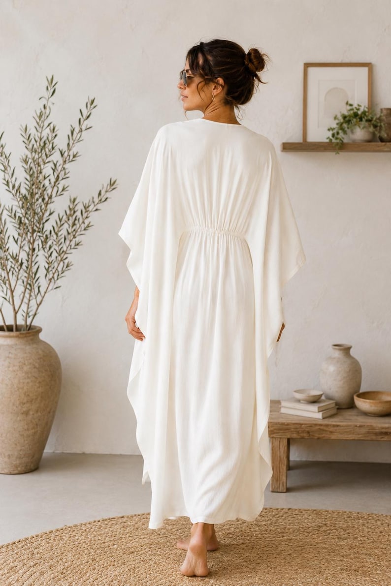 May include: A flowing, white caftan dress with a cinched waist. The dress has wide sleeves and a V-neck. The model is wearing sunglasses and has their hair up. The background includes a neutral-toned room with a wooden shelf and a large vase.