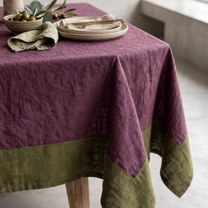 May include: A rectangular tablecloth featuring a deep plum purple centre with a contrasting olive green border. The fabric displays a subtle herringbone weave pattern throughout, draping over the edges of a wooden table with a textured appearance.