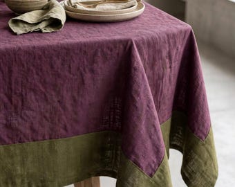 May include: A rectangular tablecloth featuring a deep plum purple center with a contrasting olive green border. The fabric displays a subtle herringbone weave pattern throughout, draping over the edges of a wooden table with a textured appearance.