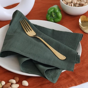 May include: A green linen napkin folded on a white plate with a gold fork. The plate is on a brown table with a green pepper and a bowl of nuts.