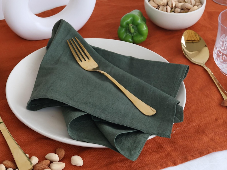 May include: A green linen napkin folded on a white plate with a gold fork. The plate is on a brown table with a green pepper and a bowl of nuts.