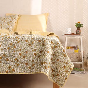 May include: A floral patterned quilt and pillow set on a bed. The quilt features a white background with brown and green floral designs. The pillows are yellow, one with stripes and the other with a matching floral pattern. The bed has a yellow border.