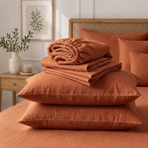 May include: A stack of solid burnt orange bedding consisting of two rectangular pillows, a folded flat sheet, and a fitted sheet with elastic edges. The fabric has a visible woven texture, and the items are arranged neatly on a bed surface.