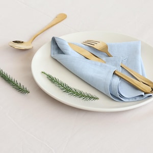May include: A white plate with a light blue linen napkin folded on top. A gold fork and knife are placed on top of the napkin. A gold spoon is on the table to the left of the plate.