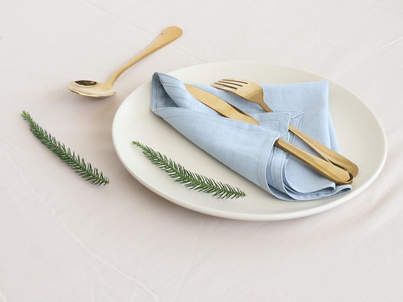 May include: A white plate with a light blue linen napkin folded on top. A gold fork and knife are placed on top of the napkin. A gold spoon is on the table to the left of the plate.