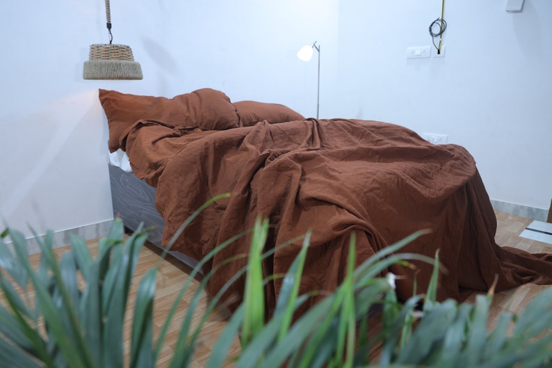 May include: A brown linen duvet cover and pillowcases are on a bed with a wooden frame. The bed is in a room with white walls and a plant in the foreground.