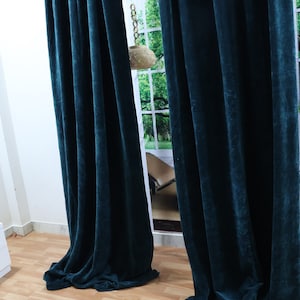 May include: Two dark teal velvet curtains hanging in a room with a wooden floor. The curtains are pulled closed, blocking out the light from the window behind them.