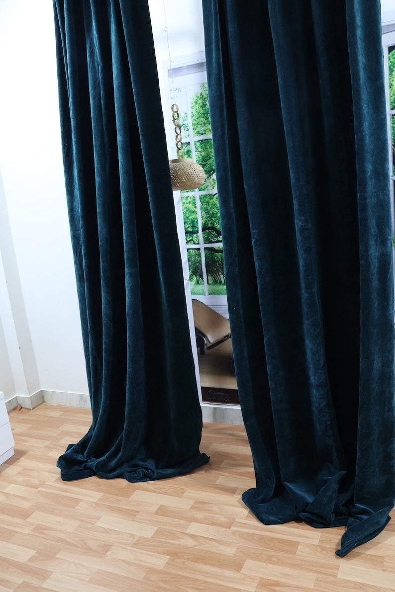 May include: Two dark teal velvet curtains hanging in a room with a wooden floor. The curtains are pulled closed, blocking out the light from the window behind them.