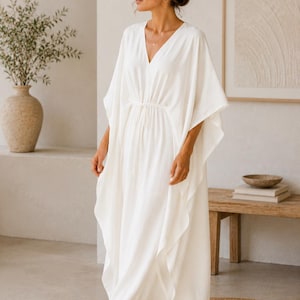 May include: A flowing white caftan dress with a V-neck and wide sleeves. The dress has a drawstring waist and falls to the ankles. The model is wearing sunglasses and standing on a round, woven rug. The background includes a vase with greenery and a wooden bench.