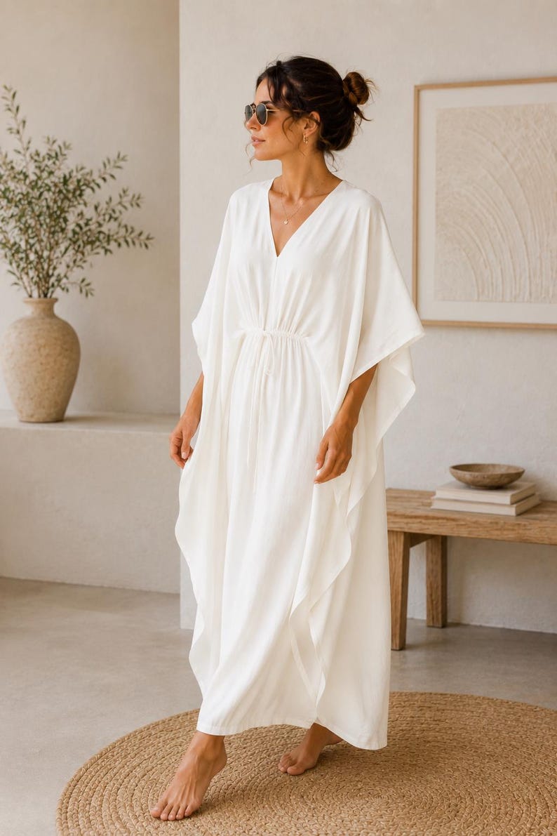 May include: A flowing white caftan dress with a V-neck and wide sleeves. The dress has a drawstring waist and falls to the ankles. The model is wearing sunglasses and standing on a round, woven rug. The background includes a vase with greenery and a wooden bench.