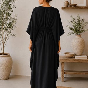 May include: A black, floor-length kaftan dress with a V-neck and wide sleeves. The dress has a gathered waist and a flowing skirt. The person is wearing sunglasses and has their hair up in a bun. The background includes plants and wooden furniture.