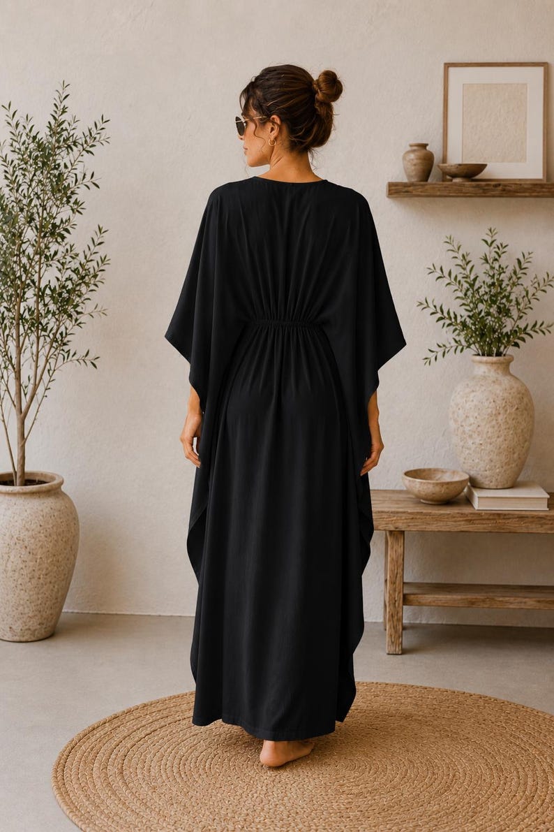 May include: A black, floor-length kaftan dress with a V-neck and wide sleeves. The dress has a gathered waist and a flowing skirt. The person is wearing sunglasses and has their hair up in a bun. The background includes plants and wooden furniture.