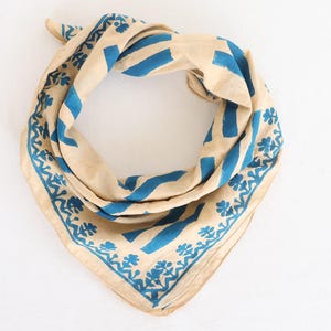 May include: A beige and blue bandana with a geometric pattern. The bandana features blue stripes and floral designs along the edges. The fabric is folded and arranged on a white surface.