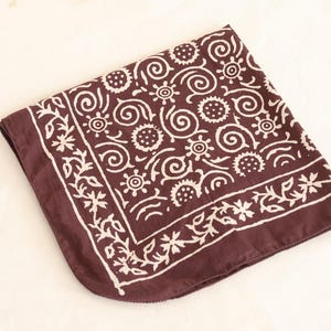 May include: A folded maroon bandana with a white floral and swirl pattern. The design includes stylized flowers, circles, and spiral shapes. The bandana is made of a soft fabric and is displayed on a light-colored surface.