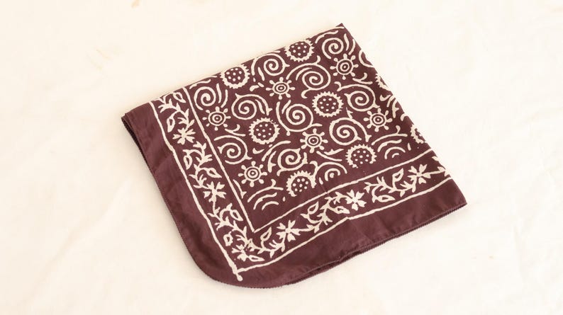 May include: A folded maroon bandana with a white floral and swirl pattern. The design includes stylized flowers, circles, and spiral shapes. The bandana is made of a soft fabric and is displayed on a light-colored surface.