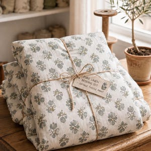 May include: A folded bedding set made of off-white linen fabric with a repeating blue and green thistle floral print. The set is tied with thin twine and features a rectangular paper tag that reads PURE LINEN BEDDING SET COTTAGE THISTLE PRINT.