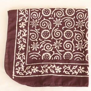 May include: A folded square textile in a deep maroon color, featuring a repeating white pattern of floral and spiral designs. The border has a floral pattern. The textile is likely a scarf or bandana.