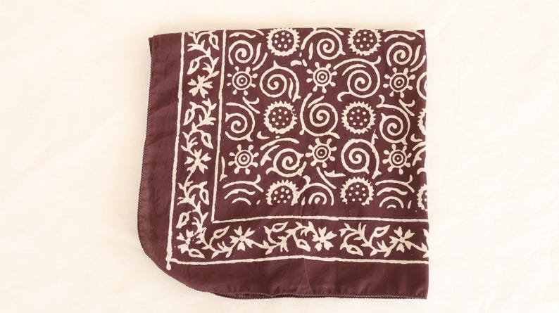 May include: A folded square textile in a deep maroon color, featuring a repeating white pattern of floral and spiral designs. The border has a floral pattern. The textile is likely a scarf or bandana.