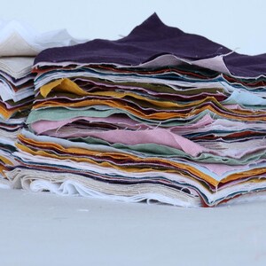 May include: A stack of fabric scraps in various colors, including white, purple, pink, yellow, and green. The fabric is folded neatly and arranged in a pile.